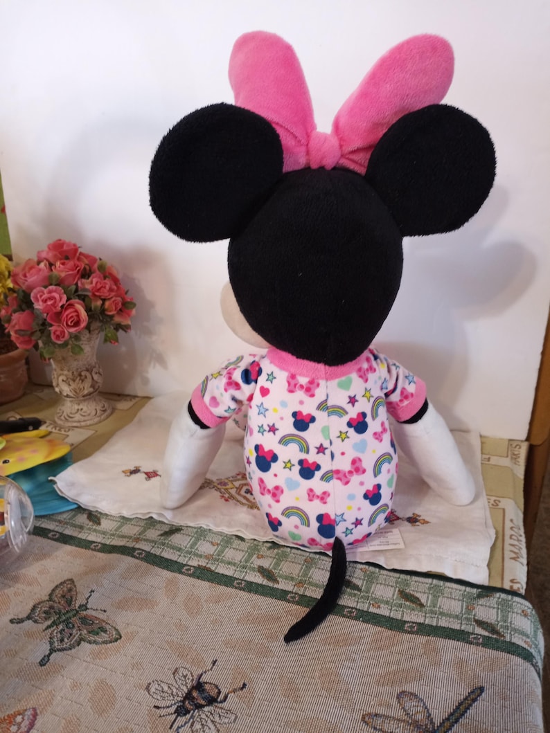 K&ouml;nnte beinhalten: Ein Pl&uuml;schtier von Minnie Mouse, das einen rosa Schlafanzug mit einem Regenbogen-, Sterne- und Herzmuster tr&auml;gt. Sie hat eine rosa Schleife auf dem Kopf und schwarze Ohren.