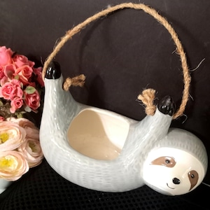 Puede incluir: Una maceta colgante de cerámica en forma de perezoso de color azul claro con un asa de cuerda. El perezoso tiene una cara blanca con ojos marrones y una expresión sonriente. El interior de la maceta es beige y las garras del perezoso son negras.