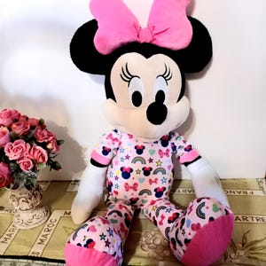 K&ouml;nnte beinhalten: Ein gro&szlig;es Pl&uuml;schtier von Minnie Mouse, das einen rosa Schlafanzug mit Regenbogen- und Sternenmuster tr&auml;gt. Sie hat eine rosa Schleife und ein breites L&auml;cheln.