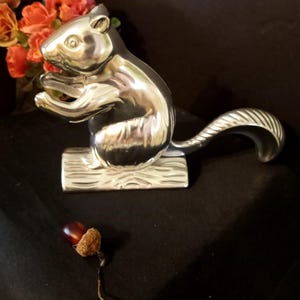 May include: A polished silver-toned metal nutcracker shaped like a squirrel sitting on a textured log base, featuring a curved tail and holding a small object. A single brown acorn with a textured cap and a thin string is positioned nearby.