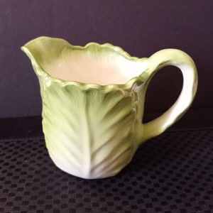 Puede incluir: Una jarra de cerámica con diseño de hoja de col. La jarra es de color verde claro con interior blanco y asa curva. La jarra tiene un borde festoneado y está sobre una superficie oscura.