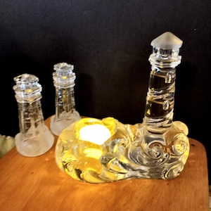 Può includere: Un portacandele a forma di faro in vetro trasparente con la parte superiore smerigliata e un design a spirale. Il portacandele ha una base con un design a spirale e contiene una candela accesa. Sono presenti anche due portacandele a forma di faro più piccoli.