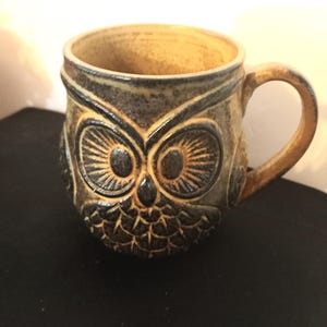 May include: A ceramic mug with an owl design. The mug is brown and tan with a detailed owl face carved into the front. The handle and rim are tan, and the owl's eyes and feathers are dark brown.