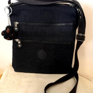 Könnte beinhalten: Eine schwarze Umhängetasche mit langem, verstellbarem Riemen, zwei horizontalen Reißverschlusstaschen auf der Vorderseite und einem runden Logo-Aufnäher. Ein kleiner, flauschiger schwarzer Affen-Schlüsselanhänger hängt am seitlichen Ring.