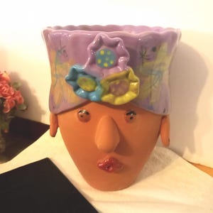 May include: A terracotta-colored ceramic planter shaped like a human head with a lavender-colored headband featuring three dimensional sculpted flowers in blue, yellow, and green. The face has painted eyes, a prominent nose, and red lips.