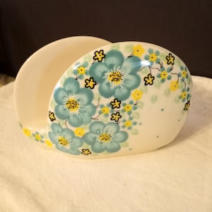 May include: A ceramic napkin holder with a curved design. The holder is white with a floral pattern of blue flowers with yellow centers and accents. The flowers are surrounded by small yellow blossoms.
