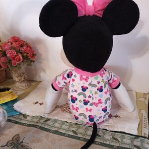 K&ouml;nnte beinhalten: Ein Pl&uuml;schtier von Minnie Mouse, das einen rosa Schlafanzug mit Regenbogen- und Sternenmuster tr&auml;gt. Sie hat eine rosa Schleife auf dem Kopf und einen schwarzen Schwanz.