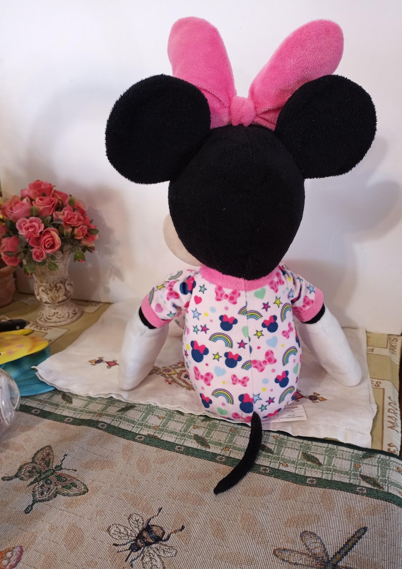 K&ouml;nnte beinhalten: Ein Pl&uuml;schtier von Minnie Mouse, das einen rosa Schlafanzug mit Regenbogen- und Sternenmuster tr&auml;gt. Sie hat eine rosa Schleife auf dem Kopf und einen schwarzen Schwanz.
