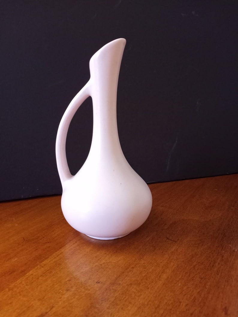 May include: A white ceramic vase with a slender neck and a rounded body. The vase has a handle on one side and a smooth, glossy finish.
