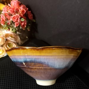 May include: A handmade ceramic bowl with a unique, wavy rim. The bowl features a gradient of colors, including brown, blue, and yellow. The base is white. The bowl is a decorative piece for the home.