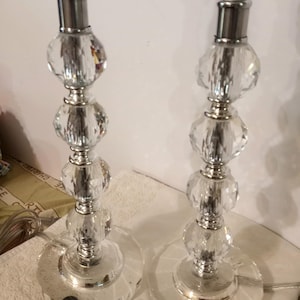 Puede incluir: Dos lámparas de mesa de cristal con detalles metálicos plateados. Cada lámpara tiene una base y un tallo compuesto por esferas de cristal facetadas. Las lámparas tienen portalámparas negros y bases redondas transparentes. Lámparas para interiores.