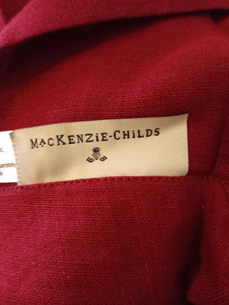 Może przedstawiać: Zbliżenie na głęboko czerwoną tkaninę z metką z napisem "MacKenzie-Childs" czarnym tekstem. Etykieta ma mały emblemat pod nazwą marki. Tkanina wydaje się być teksturowanym lnem lub podobnym materiałem.