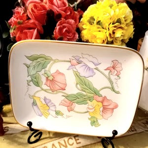 May include: A rectangular, white ceramic dish with a gold rim, featuring a floral design in pastel colors. The flowers are pink, purple, and yellow with green leaves. The dish is displayed on a black metal stand.