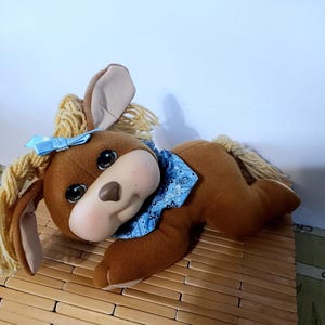 May include: A brown plush toy dog with large black eyes, a light pink nose, and a blue bandana. The dog has long, floppy ears and a light blue bow on its head. The toy is lying down on a bamboo surface.