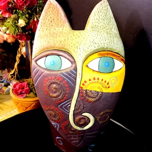 Può includere: Una maschera in ceramica a forma di gatto con un design stravagante. La maschera presenta un volto diviso con un lato nei toni del marrone e l'altro in giallo. Gli occhi sono blu e il naso ha una forma arricciata. È un'opera d'arte decorativa.