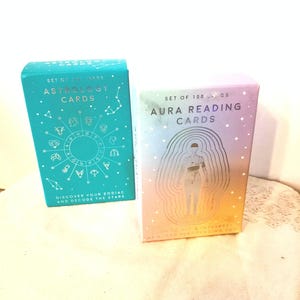 May include: Two card decks, one teal and one pastel, are displayed. The teal box reads "Astrology Cards" with zodiac symbols. The pastel box reads "Aura Reading Cards" with a human figure illustration. Both boxes are labeled "Set of 100 Cards".