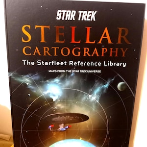 Könnte beinhalten: Ein Hardcover-Buch mit dem Titel "STAR TREK STELLAR CARTOGRAPHY" mit schwarzem Einband. Das Buch zeigt eine Weltraumszene mit einem Raumschiff und Planeten. Der Titeltext ist orange und weiß.