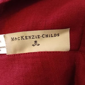 Może przedstawiać: Zbliżenie na głęboko czerwoną tkaninę z metką z napisem "MacKenzie-Childs" czarnym tekstem. Etykieta ma mały emblemat pod nazwą marki. Tkanina wydaje się być teksturowanym lnem lub podobnym materiałem.