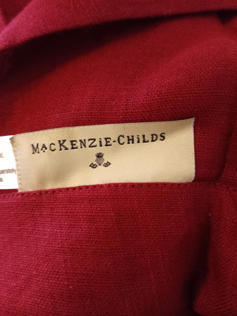 Może przedstawiać: Zbliżenie na głęboko czerwoną tkaninę z metką z napisem "MacKenzie-Childs" czarnym tekstem. Etykieta ma mały emblemat pod nazwą marki. Tkanina wydaje się być teksturowanym lnem lub podobnym materiałem.