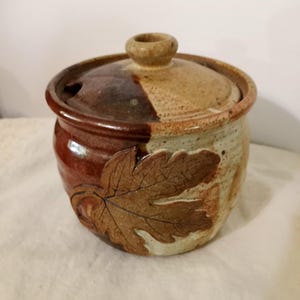 May include: A handcrafted ceramic jar with a lid. The jar is a mix of brown, tan, and cream colors, with a leaf design on the side. The lid has a small knob and a hole. The jar is likely used for storage.