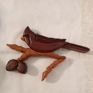May include: A wooden carving of a cardinal perched on a branch with two pine cones. The cardinal is carved from dark wood and the branch and pine cones are carved from light wood.