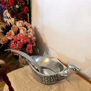 Può includere: Una ciotola da portata color argento con un design a forma di uccello, con un lungo manico e un cucchiaio all'interno. La ciotola è appoggiata su un asciugamano beige, con una composizione floreale sullo sfondo.