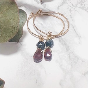 May include: Gold hoop earrings with a dangling dark green and red gemstone.