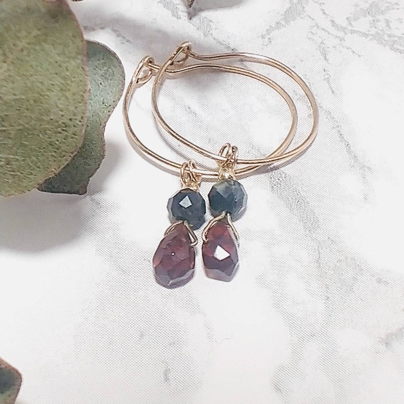 May include: Gold hoop earrings with a dangling dark green and red gemstone.