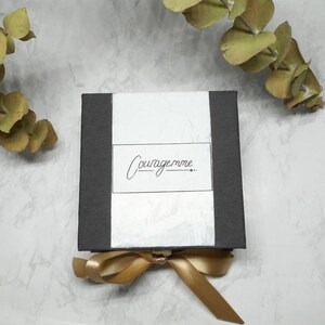 May include: A black gift box with a white label that says "Couragemme..."  The box is tied with a gold ribbon.