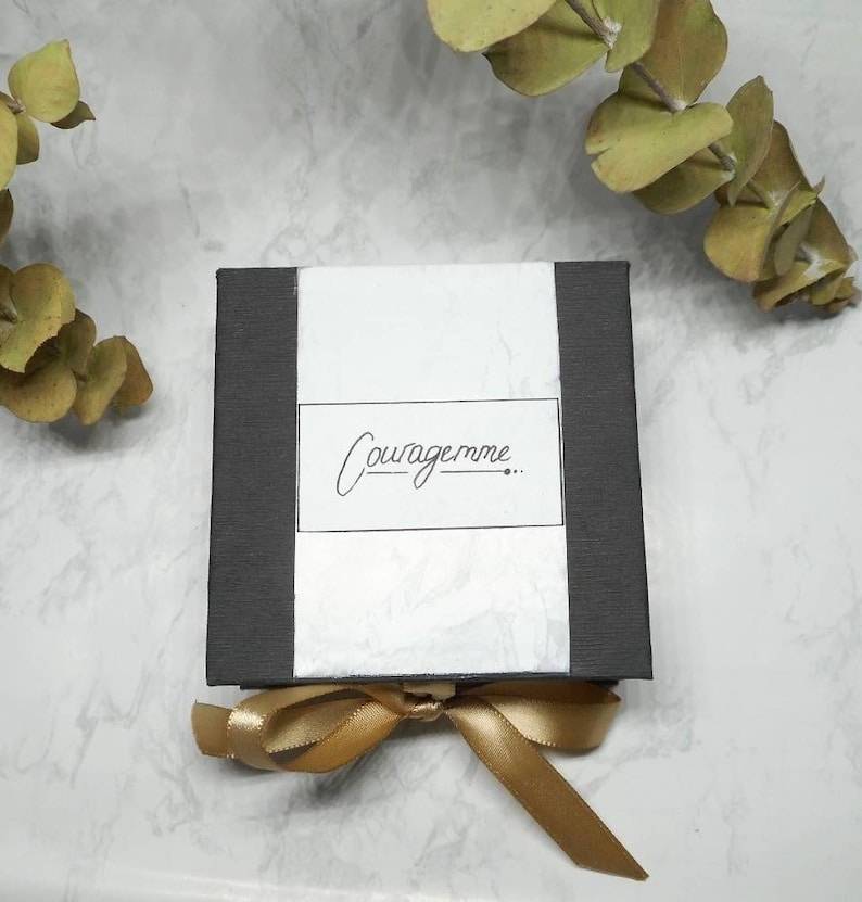 May include: A black gift box with a white label that says "Couragemme..."  The box is tied with a gold ribbon.