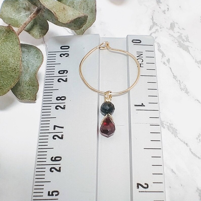 May include: Gold hoop earrings with a dangling red and black gemstone charm. The earrings are measured against a ruler, showing they are just under 1 inch in length.