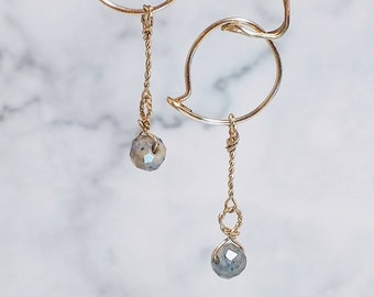 May include: A pair of gold earrings with a unique, abstract design. Each earring features a small, round, gray gemstone dangling from a delicate chain.