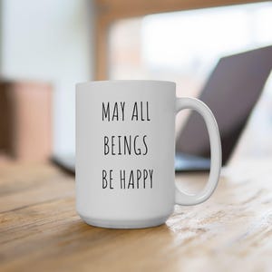 May include: White ceramic mug with the text "MAY ALL BEINGS BE HAPPY" in black. The mug has a large handle and is sitting on a wooden surface. A laptop is visible in the background.