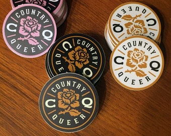 Puede incluir: Una pila de pegatinas redondas con fondo negro y un diseño de rosa dorada. El texto "COUNTRY QUEER" está escrito en un círculo alrededor de la rosa. Las pegatinas están apiladas sobre una superficie de madera.