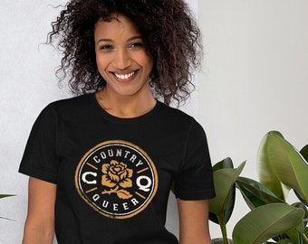 Puede incluir: Una persona que lleva una camiseta negra con un gráfico circular dorado que dice "Country Queer" con una rosa dentro del círculo.