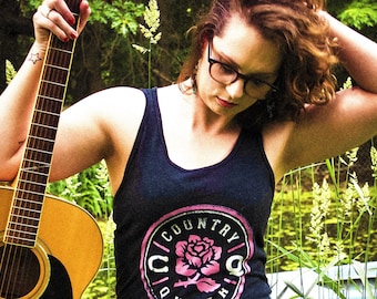 Puede incluir: Una mujer con el pelo castaño rizado lleva un top negro sin mangas con un logotipo circular rosa que dice "Country Queen" y sostiene una guitarra acústica.