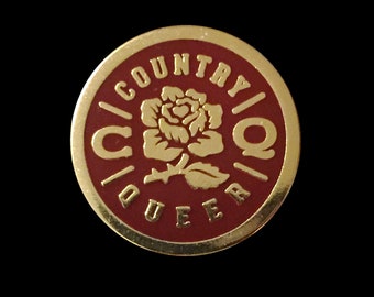 Puede incluir: Un pin de color dorado con fondo rojo. El pin presenta una rosa estilizada con tallo y hojas. Las palabras "COUNTRY" y "QUEER" están escritas alrededor de la rosa en un círculo.