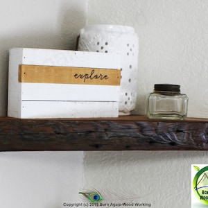 Op de afbeelding: Een rustiek houten hoekplank met een witte doos waarop "explore" staat. Er staat een glazen pot en een wit decoratief object op de plank.