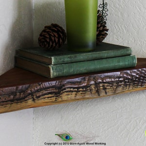 Op de afbeelding: Een rustiek houten hoekplank met een natuurlijke houten afwerking. De plank is versierd met twee groene boeken, een groene glazen vaas en twee dennenappels.