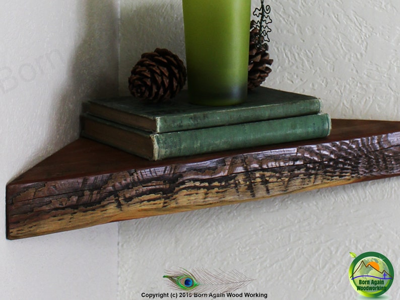 Op de afbeelding: Een rustiek houten hoekplank met een natuurlijke houten afwerking. De plank is versierd met twee groene boeken, een groene glazen vaas en twee dennenappels.