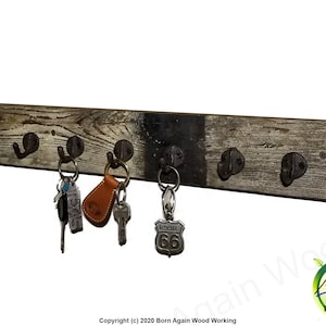 May include: A rectangular wall-mounted wooden coat rack featuring a weathered gray finish with a dark central section. It includes seven dark metal hooks, with three sets of keys hanging from the hooks, including a Route 66 keychain.