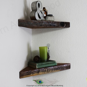 Op de afbeelding: Twee rustieke houten hoekplanken met een verweerde afwerking. De bovenste plank heeft een metalen ampersand en een glazen fles erop. De onderste plank heeft een groen glas, een dennenappel en een stapel boeken.