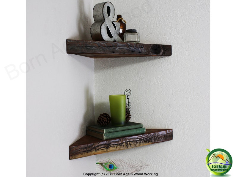Op de afbeelding: Twee rustieke houten hoekplanken met een verweerde afwerking. De bovenste plank heeft een metalen ampersand en een glazen fles erop. De onderste plank heeft een groen glas, een dennenappel en een stapel boeken.