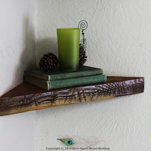 Op de afbeelding: Een rustiek houten hoekplank met een natuurlijke houten afwerking. De plank is versierd met een groen glas, twee dennenappels en een stapel boeken. De plank is gemonteerd aan een witte muur.