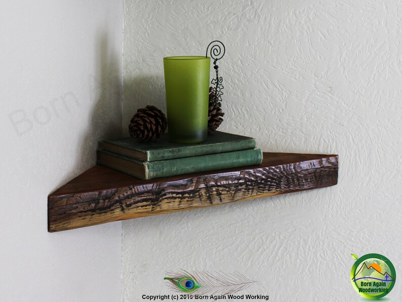 Op de afbeelding: Een rustiek houten hoekplank met een natuurlijke houten afwerking. De plank is versierd met een groen glas, twee dennenappels en een stapel boeken. De plank is gemonteerd aan een witte muur.