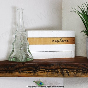 Op de afbeelding: Een wit houten bord met het woord "explore" geschreven in bruine letters op een bruine strook. Het bord staat op een houten plank met een heldere glazen fles in de vorm van de Eiffeltoren en een groene plant in een grijze pot.