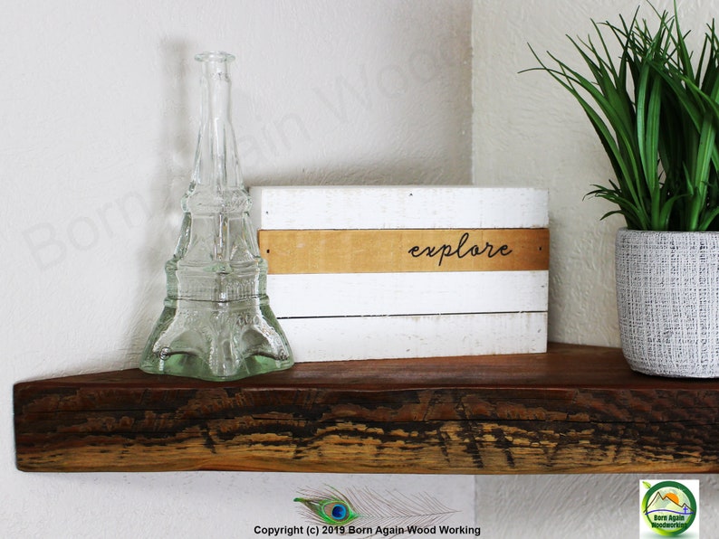 Op de afbeelding: Een wit houten bord met het woord "explore" geschreven in bruine letters op een bruine strook. Het bord staat op een houten plank met een heldere glazen fles in de vorm van de Eiffeltoren en een groene plant in een grijze pot.