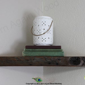 Op de afbeelding: Een rustiek houten plank met een witte decoratieve kaarsenhouder en een stapel boeken erop. De plank is gemonteerd aan een lichtbeige muur.
