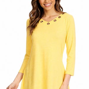May include: A bright yellow tunic top featuring three-quarter length sleeves and a V-neckline accented with metallic grommets along the collar. The garment has a flared silhouette that extends past the waistline, paired with dark blue denim jeans.