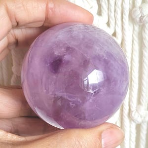 May include: A polished, spherical amethyst gemstone featuring translucent light purple hues with darker violet inclusions and natural internal veining throughout the smooth, rounded surface of the crystal ball.